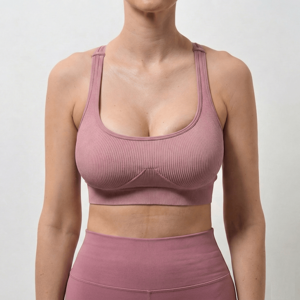 A headless, square clinical before photograph (1:1 ratio) of a white woman exhibiting severe, dramatic breast asymmetry and drooping  Her right breast (viewer's left) is significantly larger and sits noticeably lower than her refined left breast  She is preparing for a complex combined Breast Augmentation & Lift (Mastopexy) managed by Dina Elite for GCC patients, wearing a pink athletic outfit against a sterile white background 