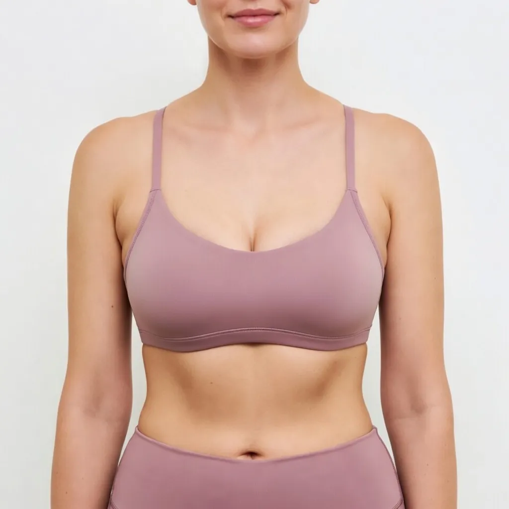 A headless, square clinical after photograph (1:1 ratio) of the same white woman, now exhibiting a restored, perky, and significantly augmented breast appearance following an Elite Breast Augmentation & Lift procedure managed by Dina Elite for GCC patients  Her breasts are symmetrical and uplifted, wearing the pink athletic outfit against the sterile white background 