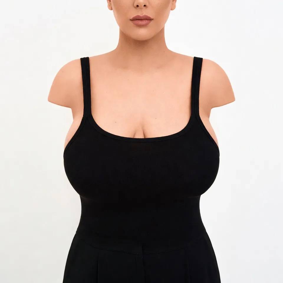 A detailed headless clinical before photograph of a Middle Eastern woman exhibiting macrosomia and breast ptosis (sagging), preparing for Elite Breast Reduction surgery managed by Dina Elite for patients from the Gulf  She is wearing a black ribbed camisole and wide leg trousers, maintaining a central standing pose on a white background 