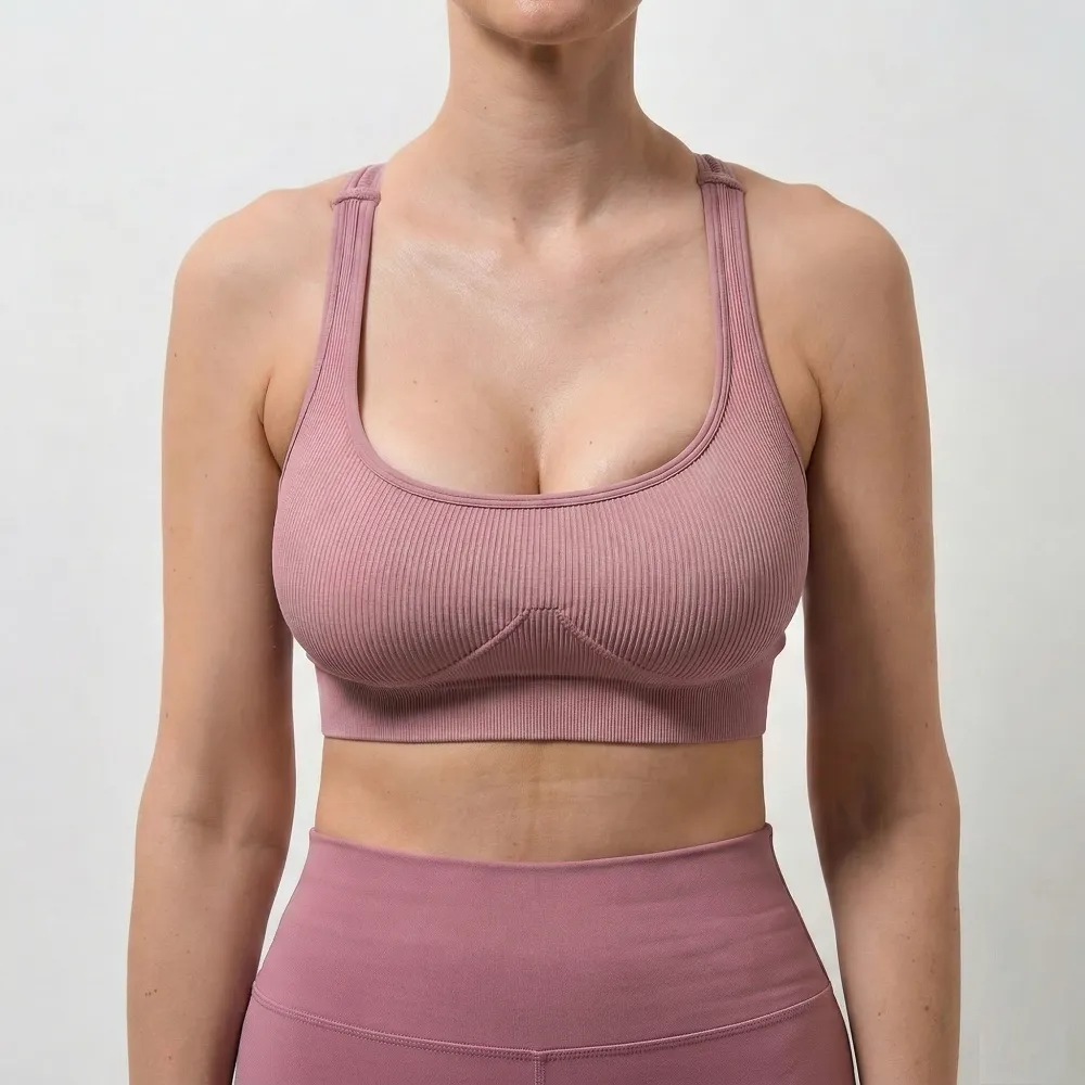 A headless, square clinical before photograph (1:1 ratio) of a white woman exhibiting severe, dramatic breast asymmetry and drooping  Her right breast (viewer's left) is significantly larger and sits noticeably lower than her refined left breast  She is preparing for a complex combined Breast Augmentation & Lift (Mastopexy) managed by Dina Elite for GCC patients, wearing a pink athletic outfit against a sterile white background 